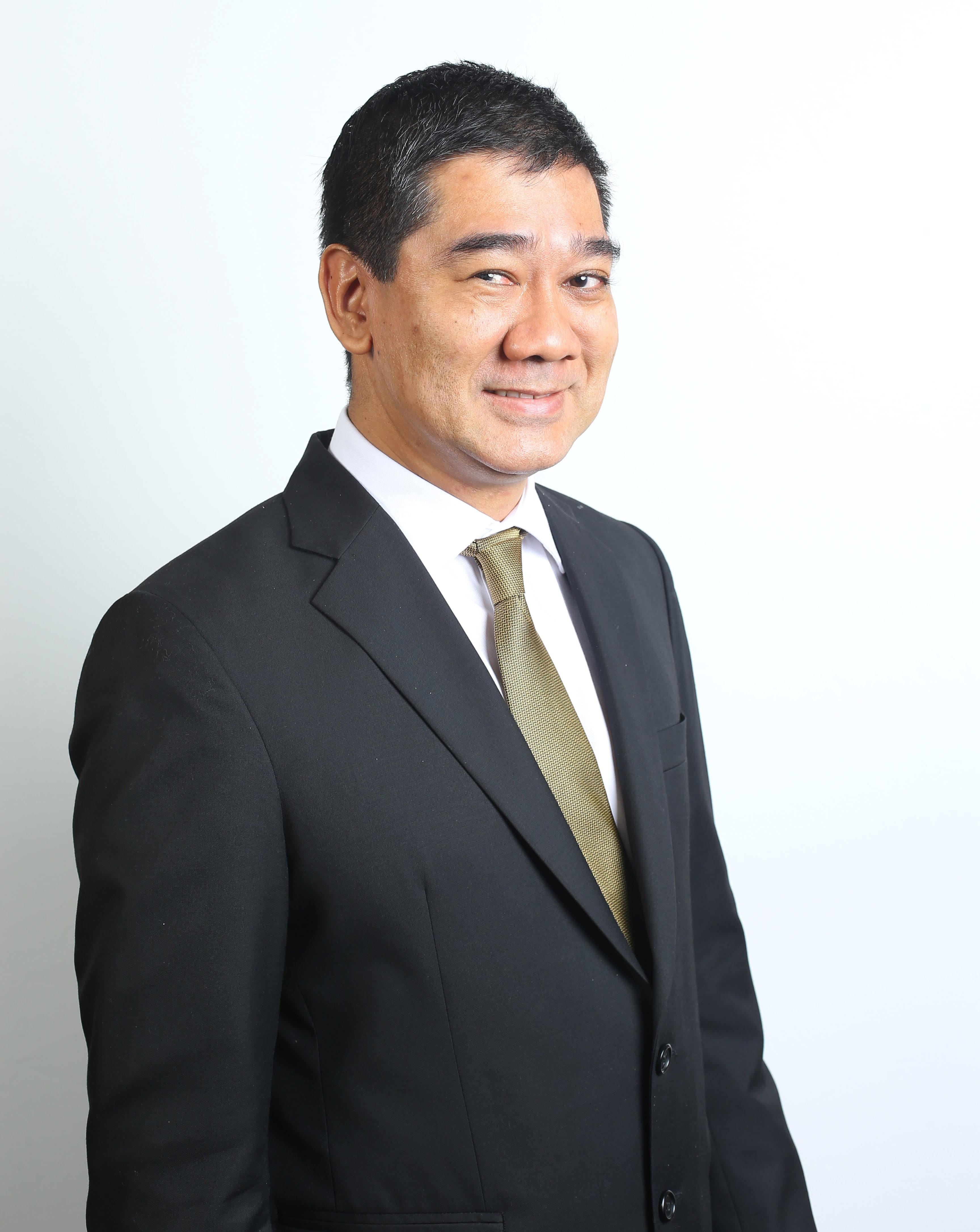 Man in black suit, white shirt, and gold patterned tie smiles against a white backdrop.